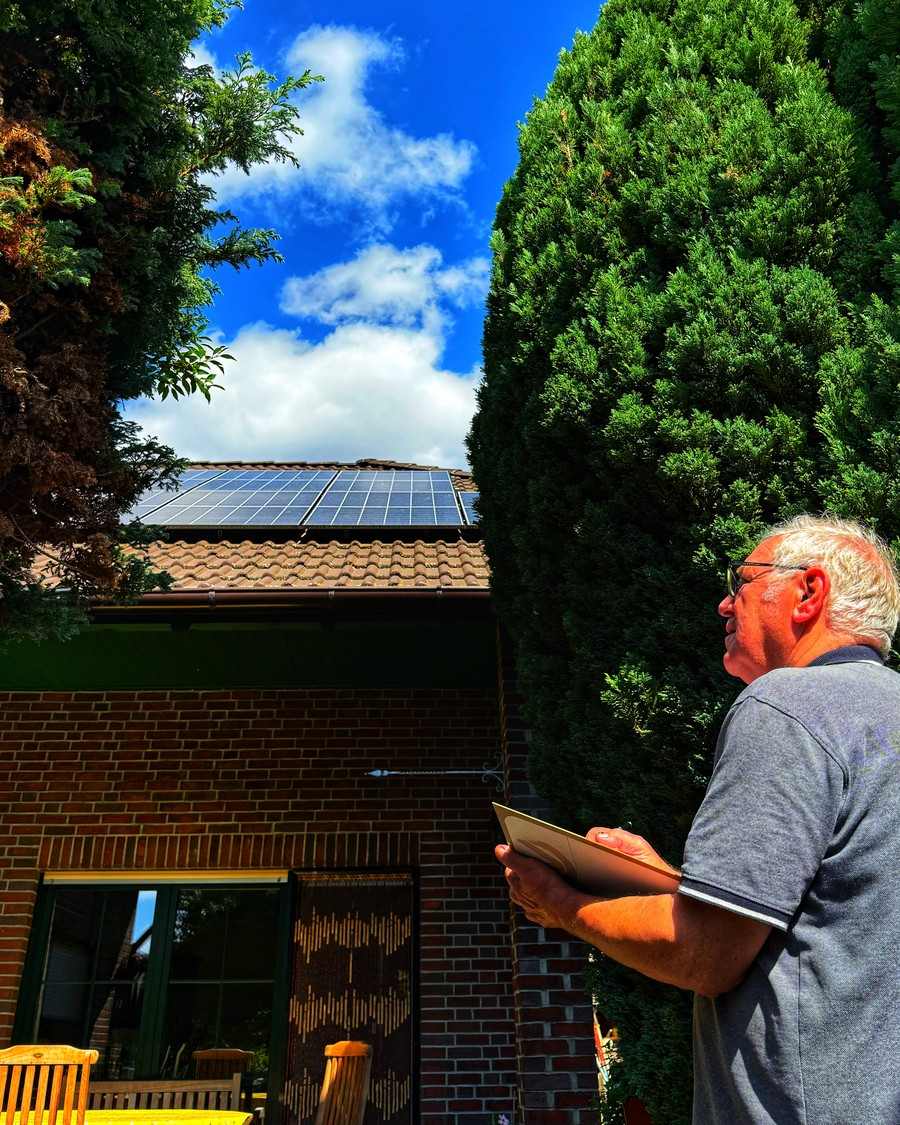 Ein Mann mit Klemmbrett blickt zu einem Ziegeldach mit Solaranlage hoch, umgeben von hohen Thuja-Hecken